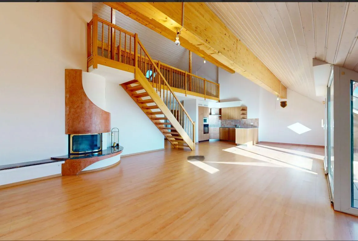 Grand séjour en duplex avec mezzanine et escalier en bois, cheminée maçonnée et cuisine ouverte.